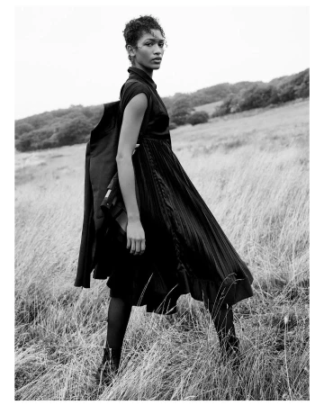 Monochrome fashion portrait of a model in a pleated black dress standing in a tall grass meadow by Scott Trindle