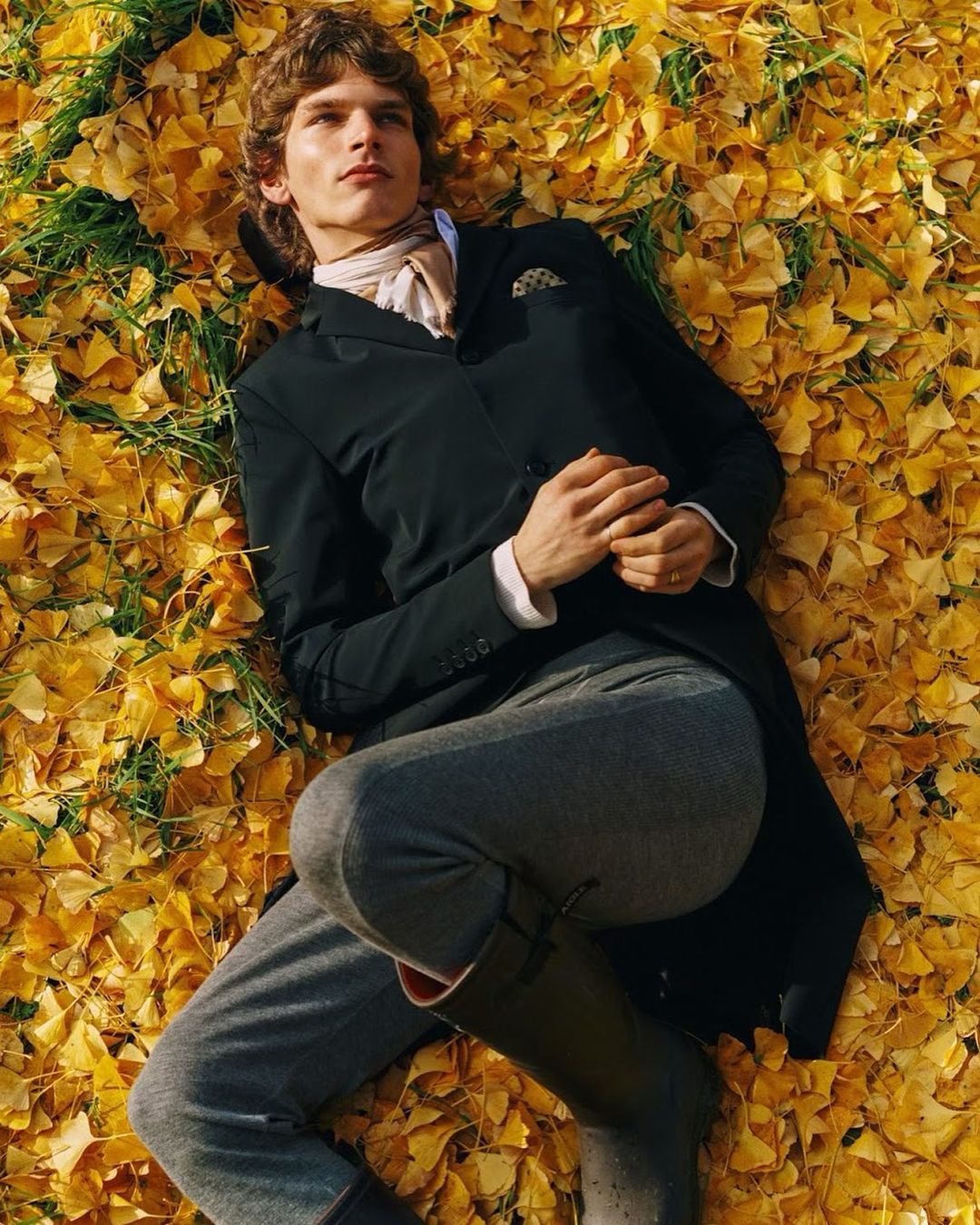 A young male model in a black jacket lies on a bed of vibrant yellow ginkgo leaves, showcasing outdoor fashion in an autumnal scene.