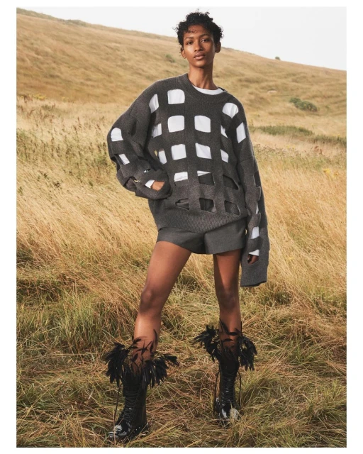 A model stands in a field wearing a grey sweater with holes, shorts, and feathered boots, against a grassy hillside backdrop.