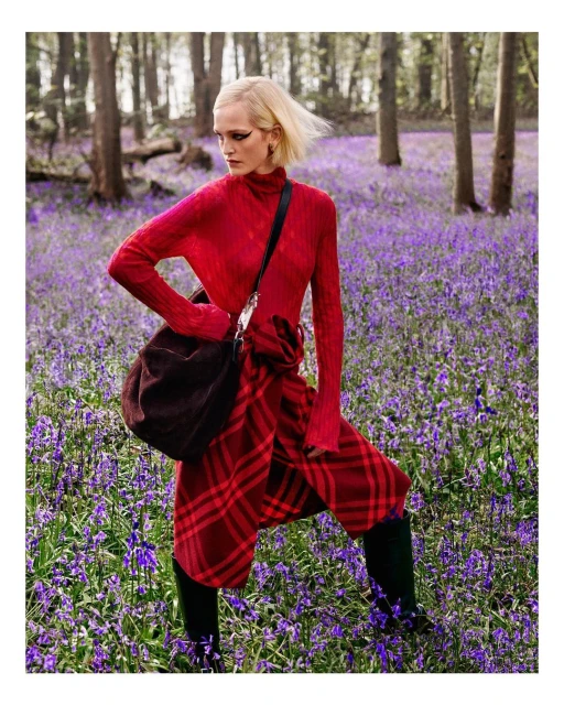 Fashion editorial shot of a blonde woman posing in a red outfit with a brown bag, standing in a woodland area filled with purple flowers.