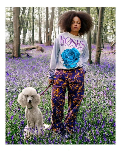 Adult woman with afro hairstyle in floral pants, standing in a bluebell field with a white standard poodle dog on leash, gazing at camera.