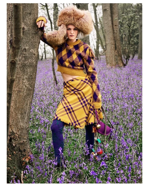 A fair-skinned woman with a large fur hat poses in a purple and yellow patterned sweater and skirt in a field of purple flowers.