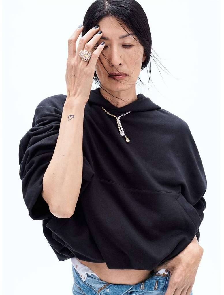 Elegant woman in a black hoodie and casual jeans, adorned with diamond jewelry, poses against a bright white backdrop in a fashion shot.