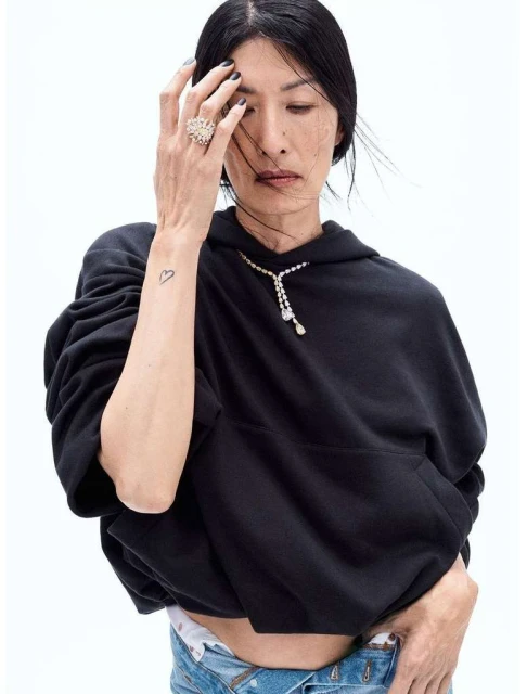 Elegant woman in a black hoodie and casual jeans, adorned with diamond jewelry, poses against a bright white backdrop in a fashion shot.