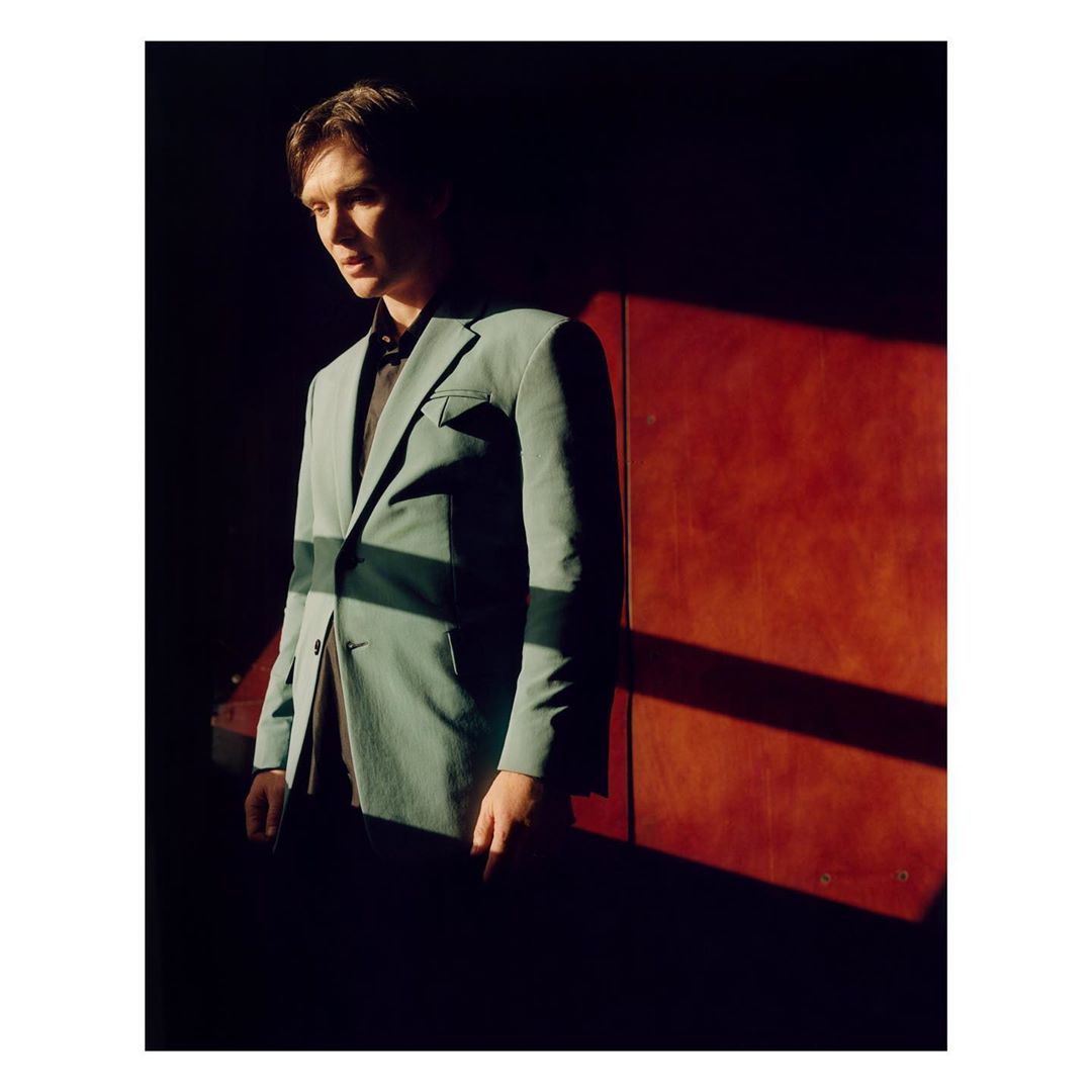 Portrait of a young man wearing a teal suit and black shirt, standing indoors in dramatic lighting casting a strong shadow on the wall.