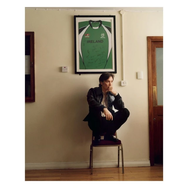 Adult male sits on a chair indoors wearing a leather jacket and dark pants. An Ireland jersey hangs on the wall.