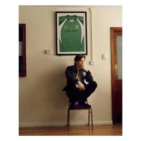 Adult male sits on a chair indoors wearing a leather jacket and dark pants. An Ireland jersey hangs on the wall.