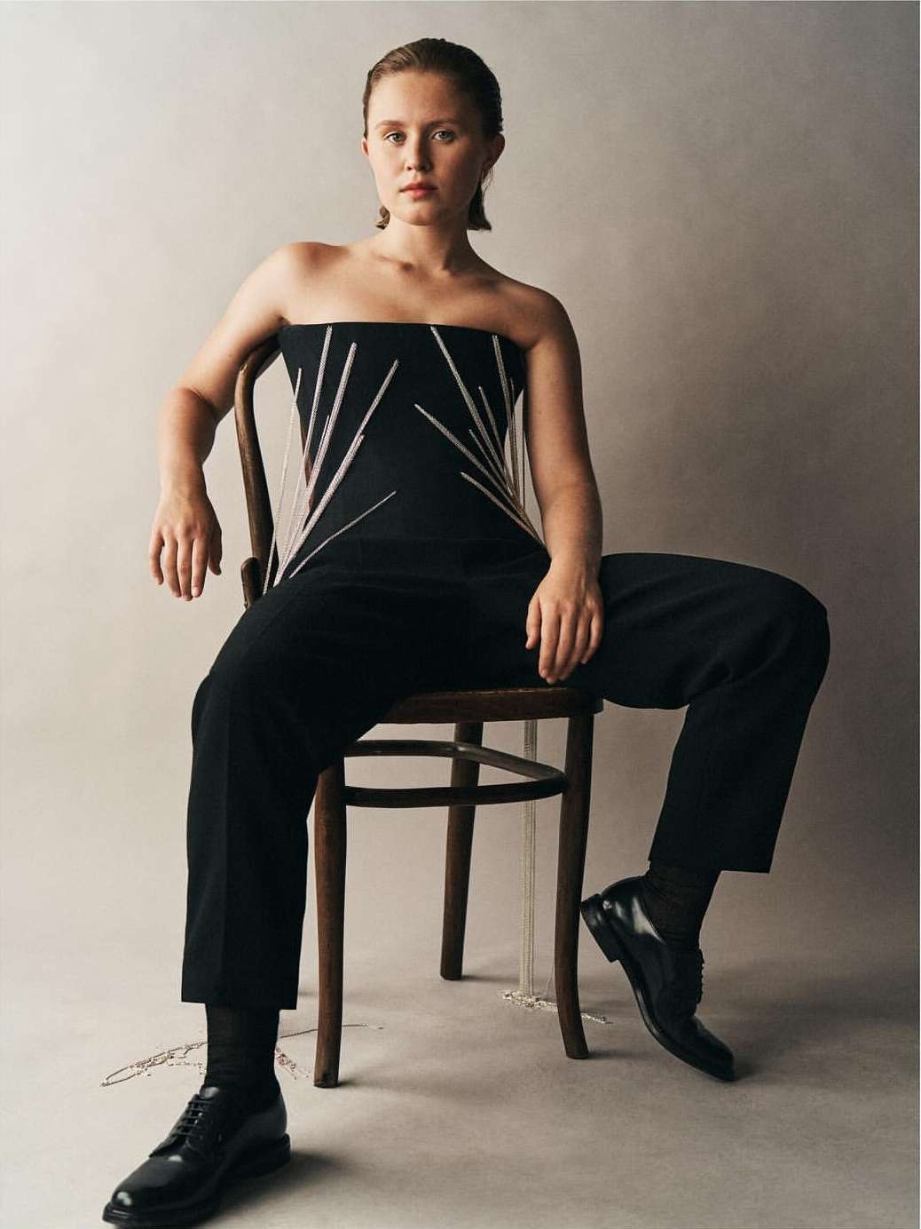 Blonde woman in a black strapless top and black pants sitting on a wooden chair in a studio, posing for a fashion shot.