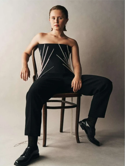 Blonde woman in a black strapless top and black pants sitting on a wooden chair in a studio, posing for a fashion shot.
