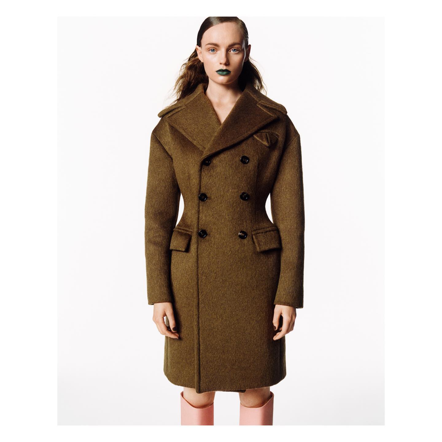 Fashion editorial shot of a woman wearing an olive-green double-breasted wool coat, green lipstick, and light-pink boots against white backdrop.