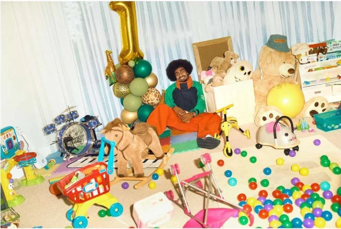 A man with an afro sits amid a sea of colorful toys and a gold balloon, in a playful, indoor scene suggesting a first birthday celebration.
