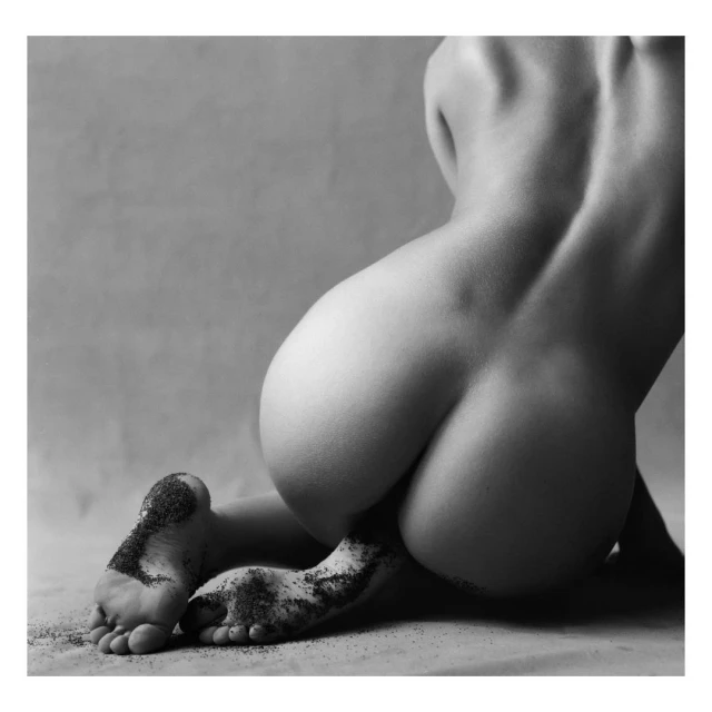 Black and white nude photo of the back of a woman kneeling, facing away with dark glitter or sand on her feet in a studio setting.