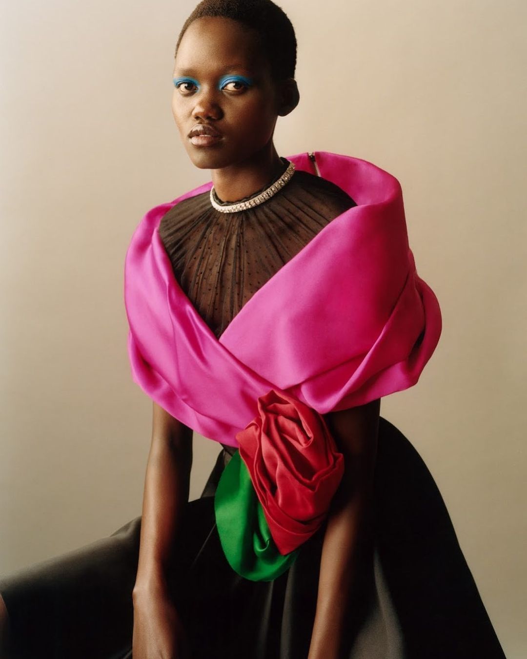 Fashion shot of an African female adult with blue eyeshadow and a pink garment with a large floral detail against a neutral backdrop.