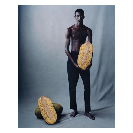 Studio shot of a Black male model holding a halved jackfruit, with additional jackfruit on the gray background, fashion-forward art.