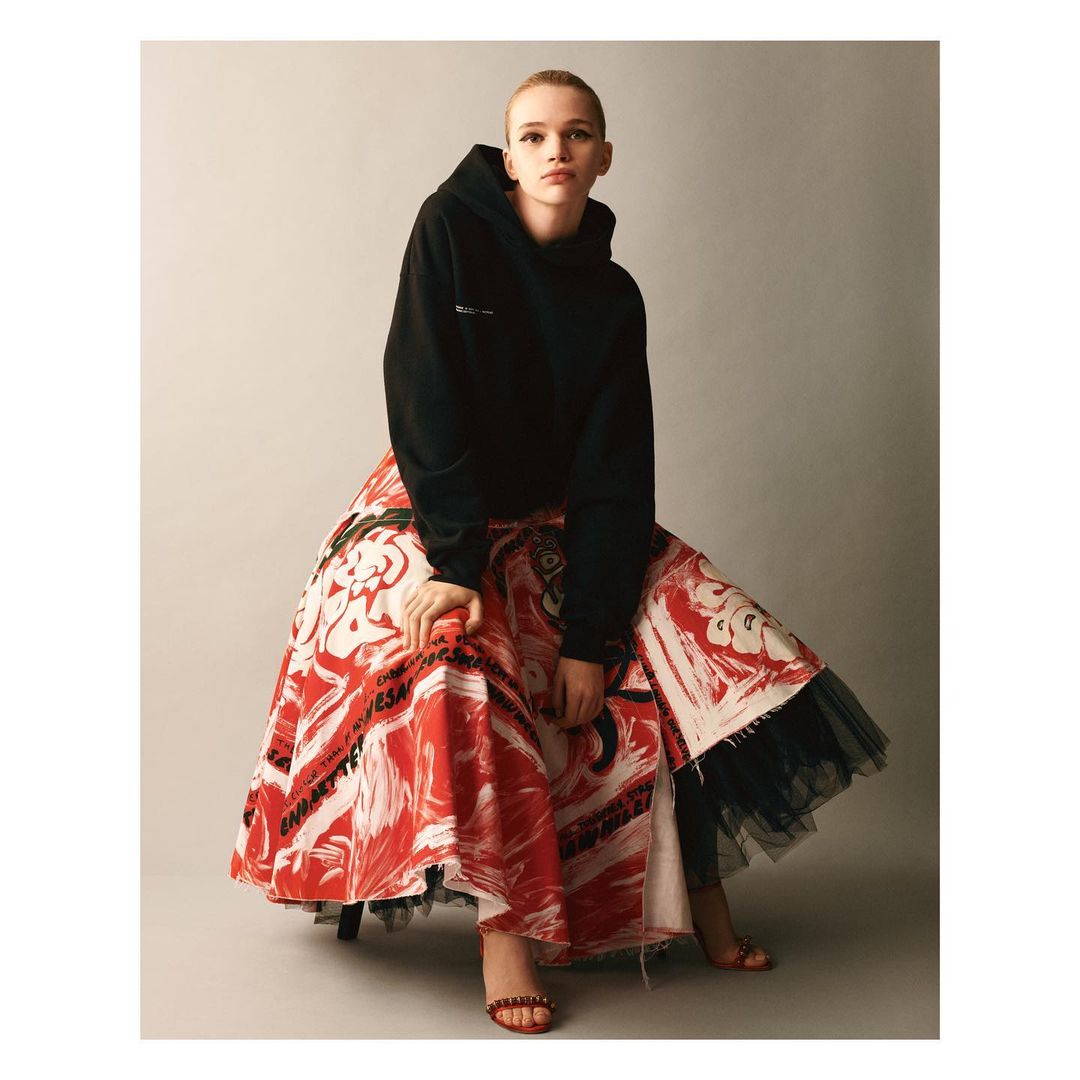 Adult female posing in a photo studio wearing a black hooded sweatshirt, long patterned skirt, and sandals, against a neutral backdrop.