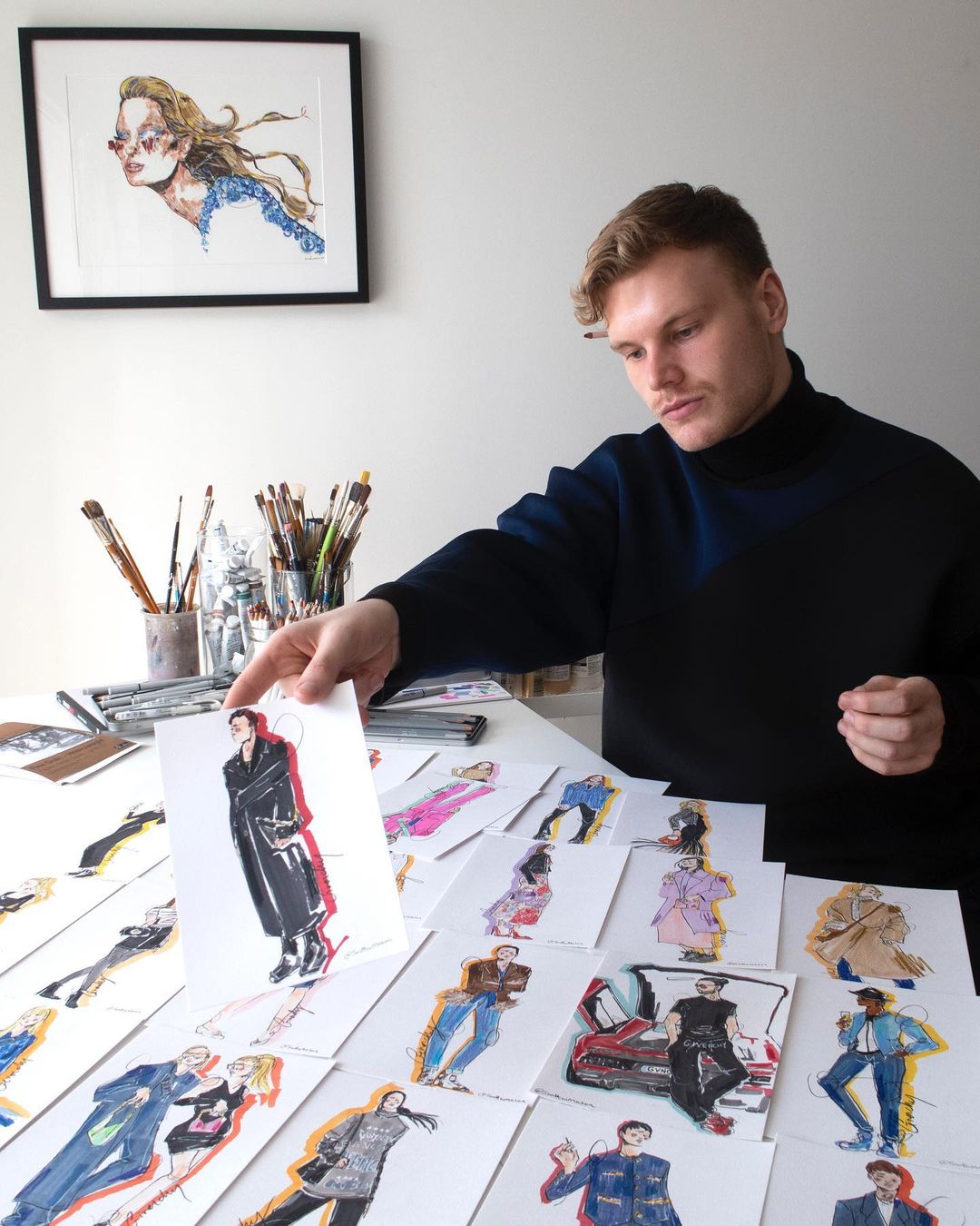 Fashion designer displaying a collection of fashion illustration drawings on a white table in a bright indoor setting, wearing black top.