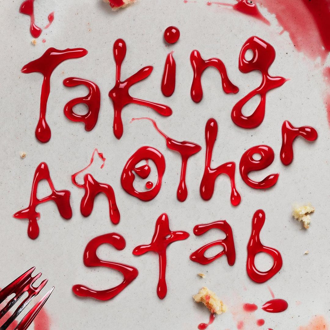Overhead shot of the words 'Taking Another Stab' written in red sauce, with cake crumbs and a fork on a neutral-toned surface.