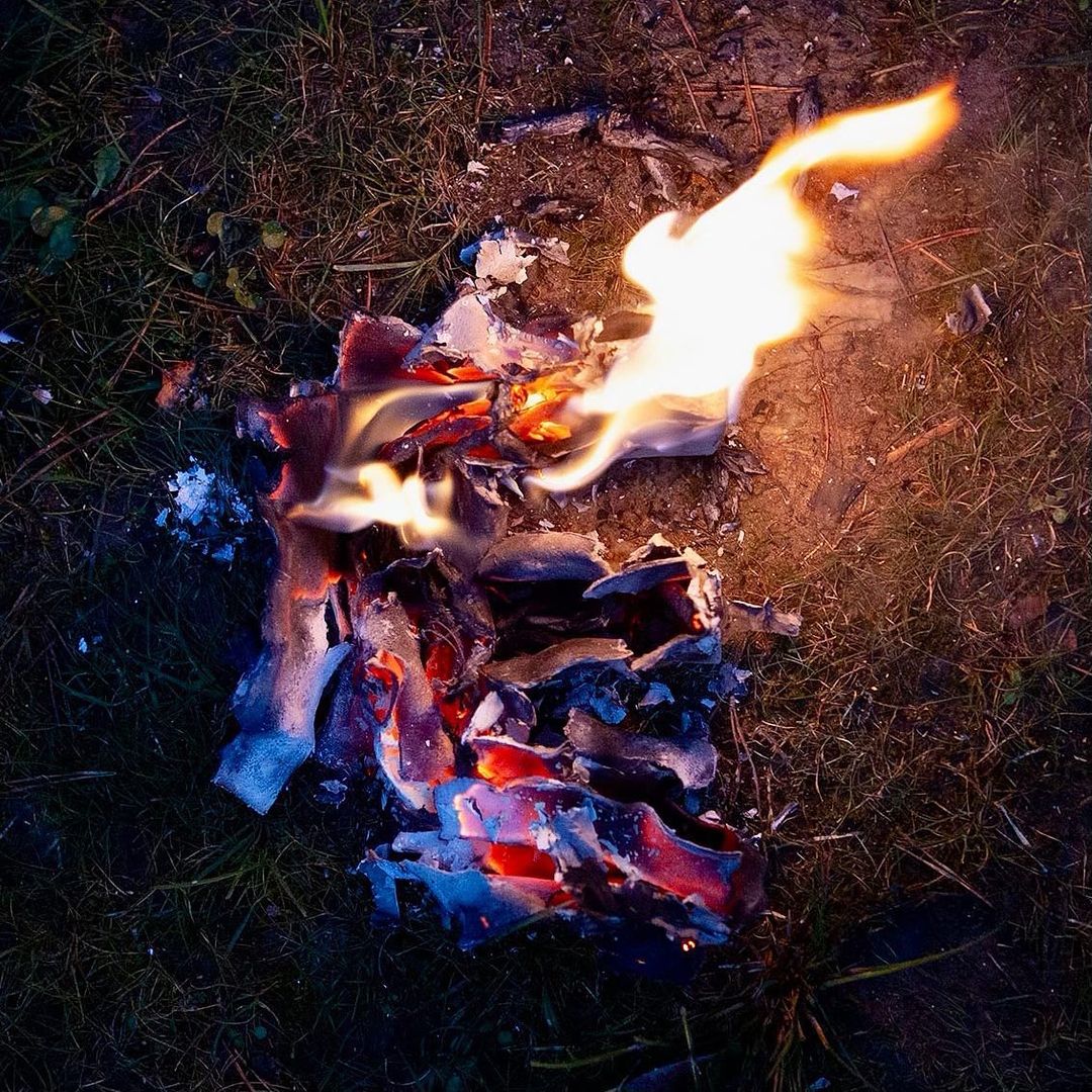Burning paper on grass ground. Flames and ashes visible in dim, outdoor light. Dark environmental setting with fire as the main subject.