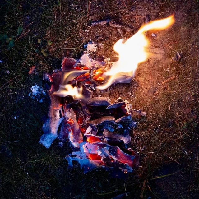 Burning paper on grass ground. Flames and ashes visible in dim, outdoor light. Dark environmental setting with fire as the main subject.