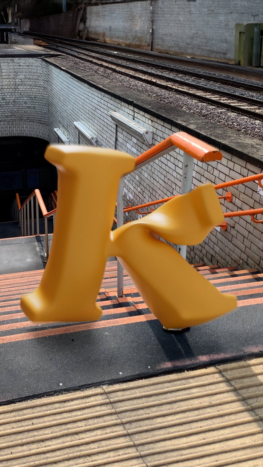 Artistic image featuring an oversized yellow letter 'K' at a train station, with railway tracks and stairs in the backdrop.