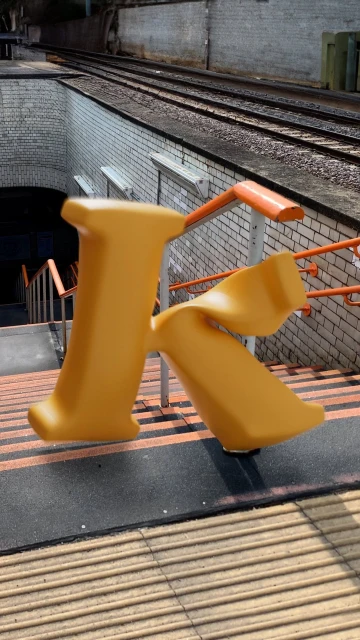Artistic image featuring an oversized yellow letter 'K' at a train station, with railway tracks and stairs in the backdrop.