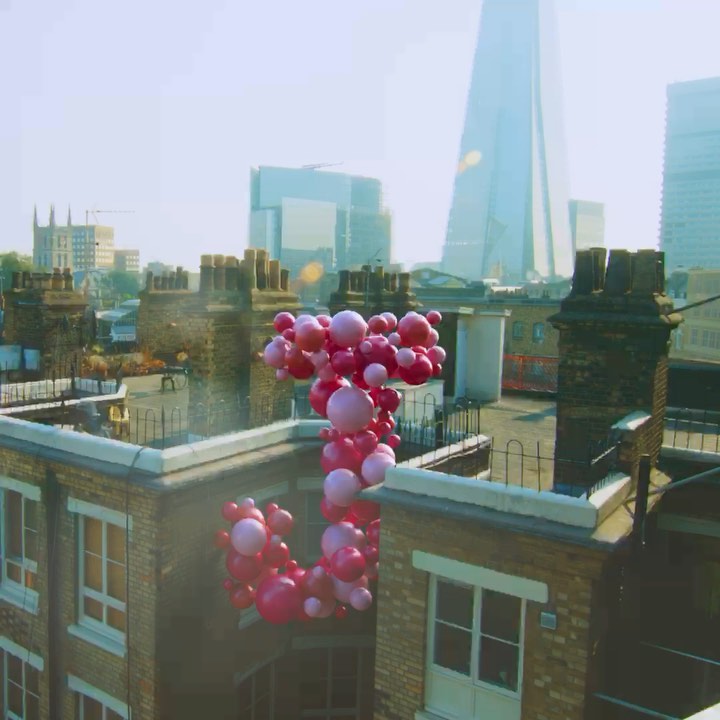 Pink balloons float above rooftops in an urban setting, featuring chimney stacks and modern architecture in the background on a sunny day.