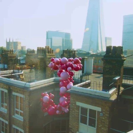 Pink balloons float above rooftops in an urban setting, featuring chimney stacks and modern architecture in the background on a sunny day.
