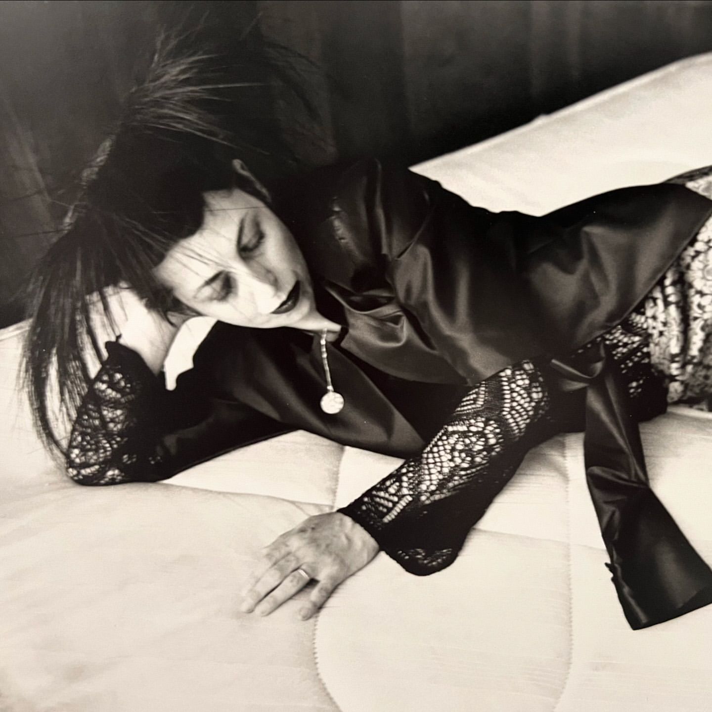 Monochrome portrait of an adult woman lying on a bed, wearing a black satin jacket and lace-detailed sleeves, captured indoors.
