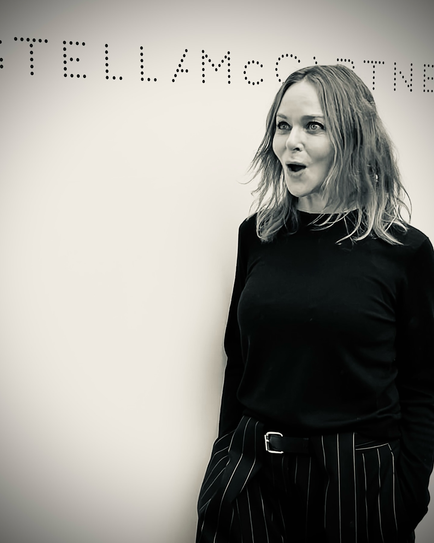 Black and white portrait of a fair-skinned adult woman with light brown hair, wearing black top and striped pants, with a surprised facial expression.