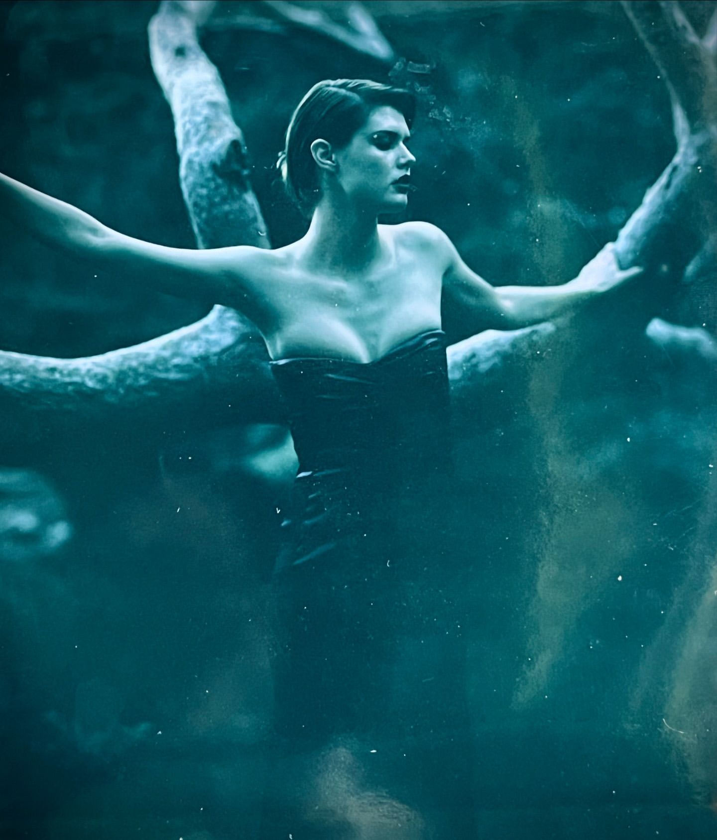 A striking portrait of an adult female in a black corset dress, surrounded by gnarled tree branches against a hazy, moody, teal background.