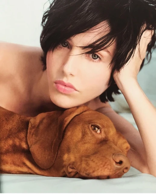 Adult woman with short dark hair rests head on Vizsla dog with amber eyes lying on white surface in a close-up portrait view.