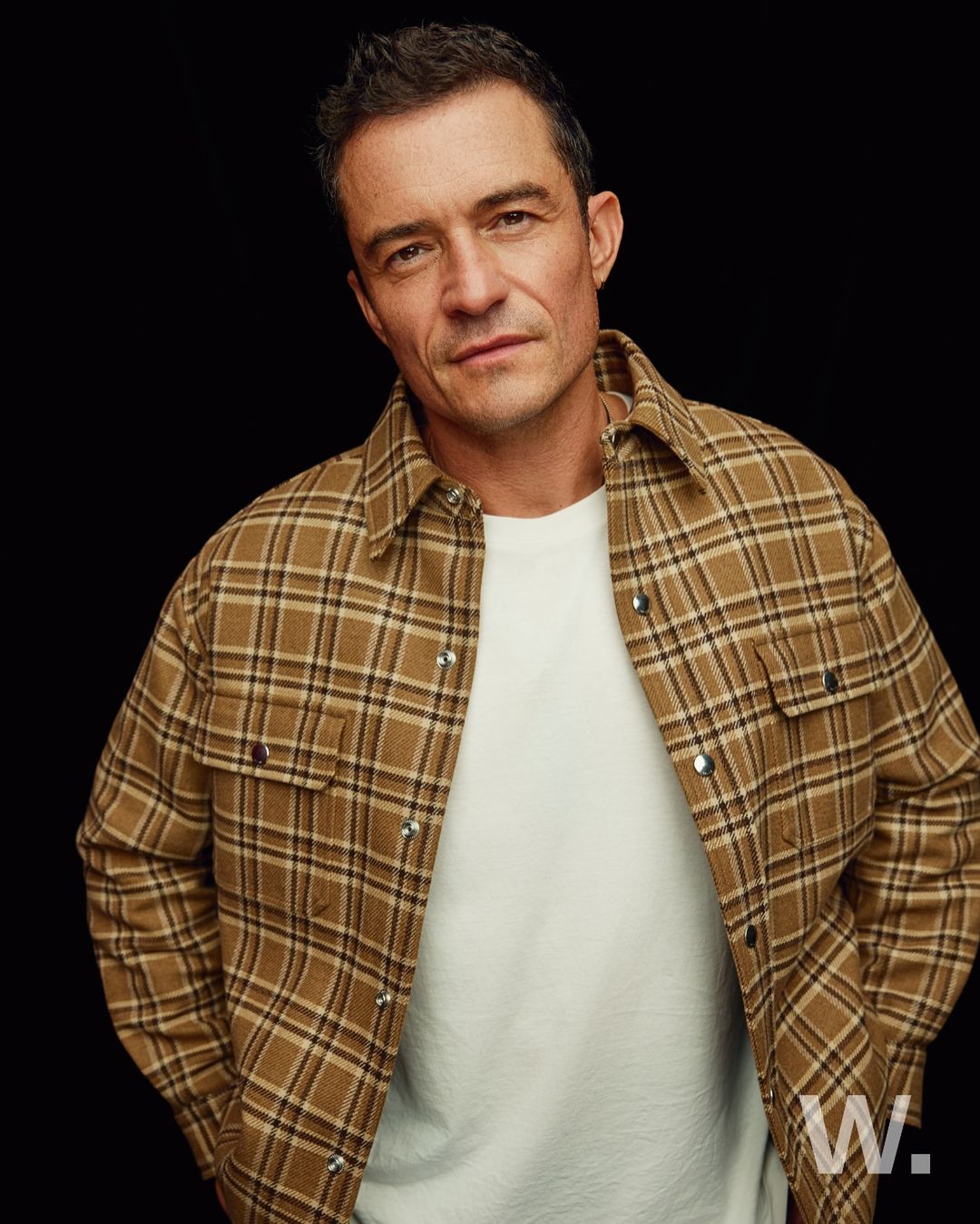 Portrait of Orlando Bloom wearing a tan plaid flannel shirt over a white t-shirt in a studio with a dark background.
