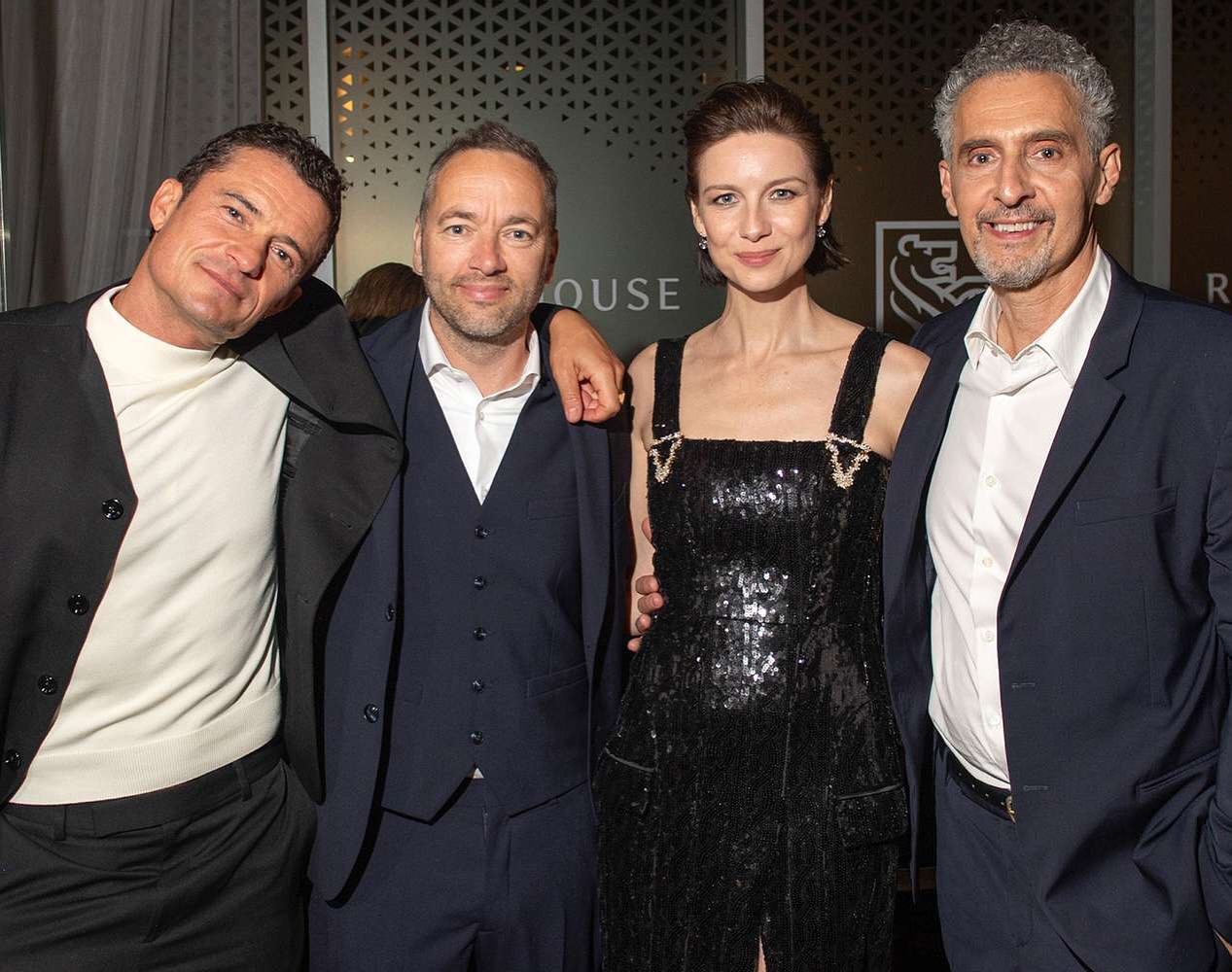 Group of four people posing in formalwear at an indoor event, smiling, with two men in dark suits, one in a sequined dress, and one in black and white.