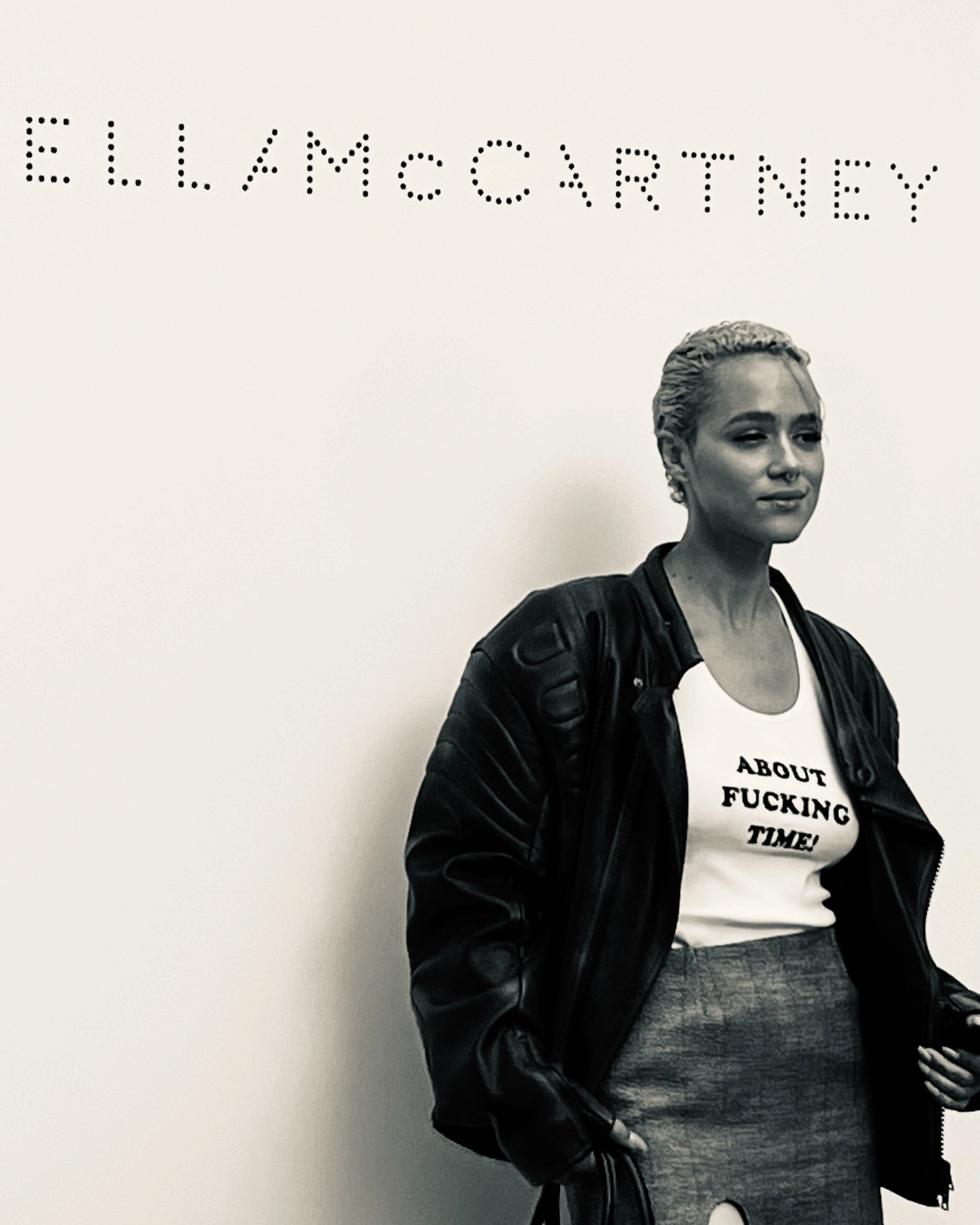 Monochrome portrait of a woman in a jacket and skirt, standing in front of a Stella McCartney logo, fashion-forward style.