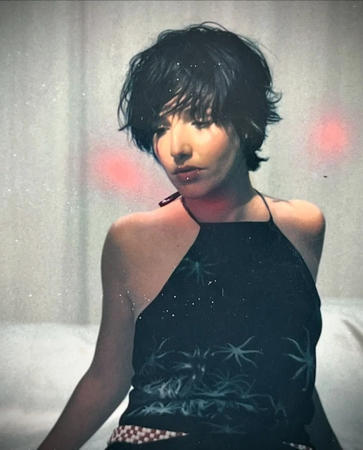 Portrait of a young adult woman with a dark halter top featuring star-like patterns, standing against a light background with diffused lighting.