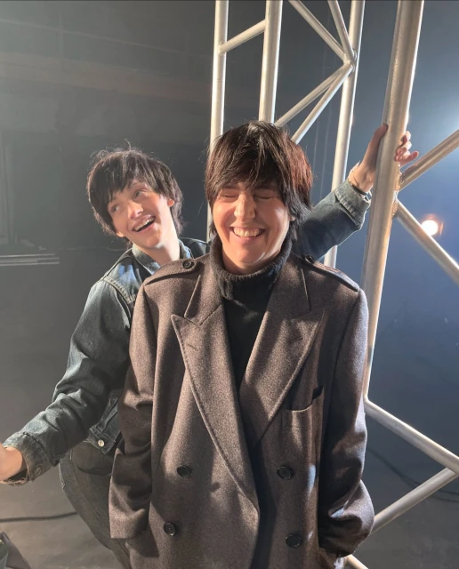 Two smiling adults pose indoors next to a metal truss. One wears a grey overcoat, the other a denim jacket in medium eye-level shot.