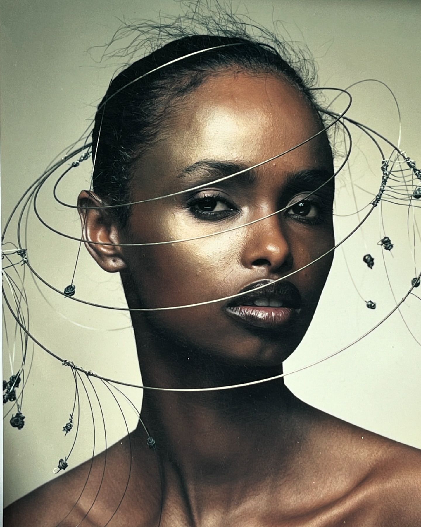 Close-up studio shot of an adult woman with dark skin and dark hair, posing with a modern art halo-like wire headpiece, against a light background.