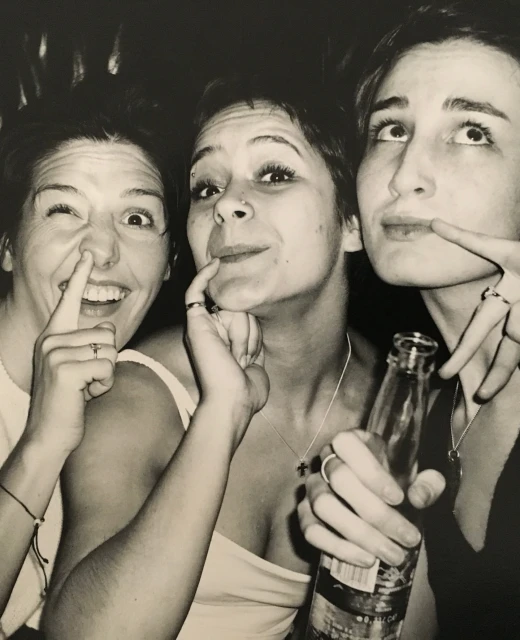 Three young women pose playfully indoors with a bottle, creating a fun, casual snapshot in monochrome tones with a dark background.