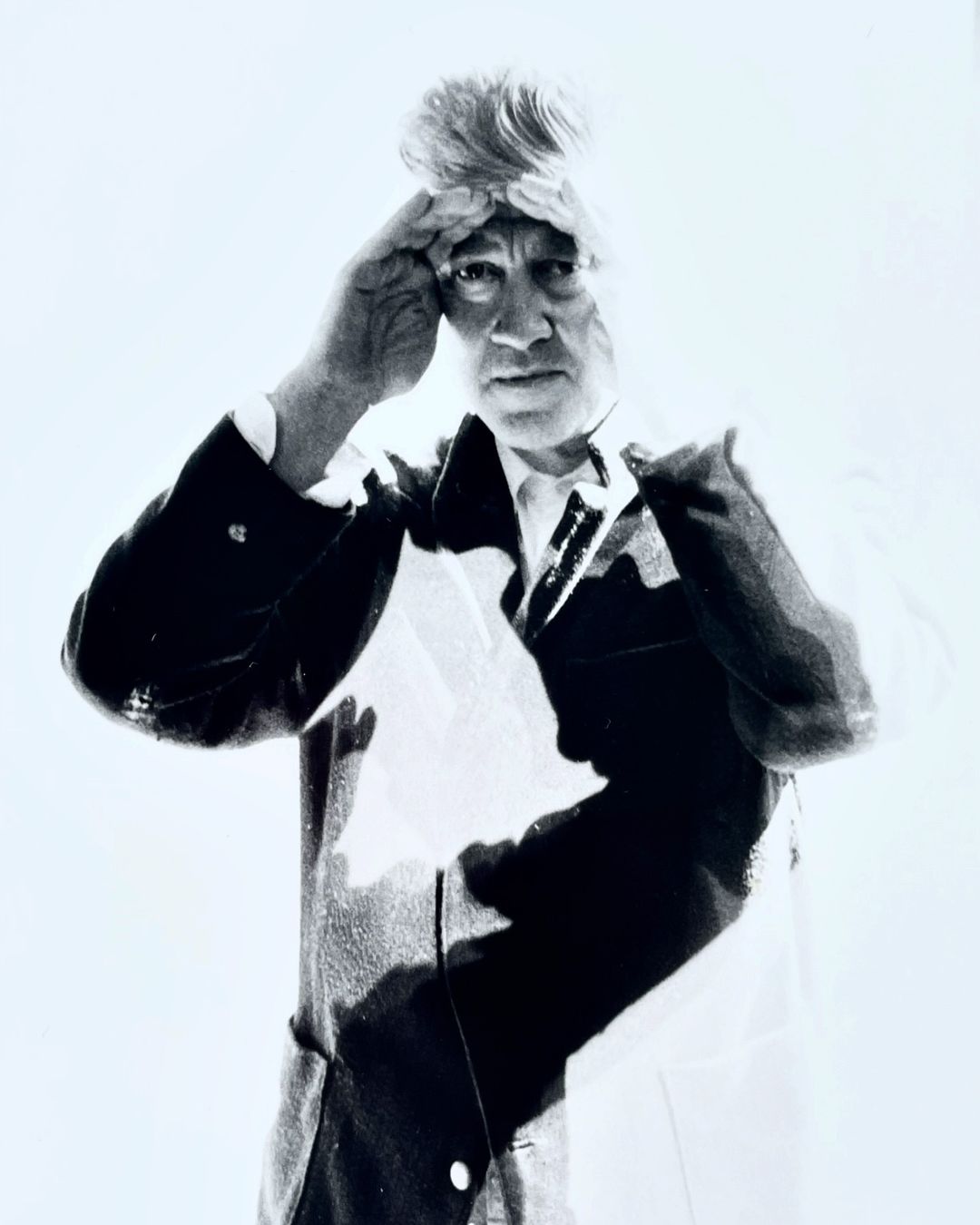 Monochrome portrait of an adult man with hand raised to his head, wearing a blazer against a bright white background in a studio setting.