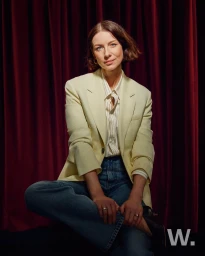 Woman with short brown hair wearing a light yellow blazer and blue jeans seated against a dark red curtain.