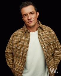 Portrait of Orlando Bloom wearing a brown plaid shirt and white t shirt against a black background
