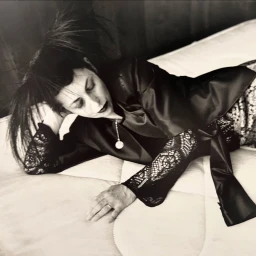 Monochrome portrait of a woman reclining on a mattress wearing a dark satin jacket with lacy patterned sleeves.