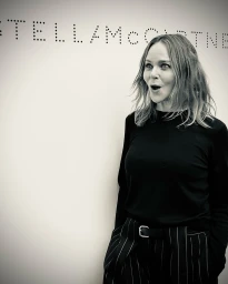 Monochrome portrait of Stella McCartney with a surprised expression wearing a dark top against a white wall.