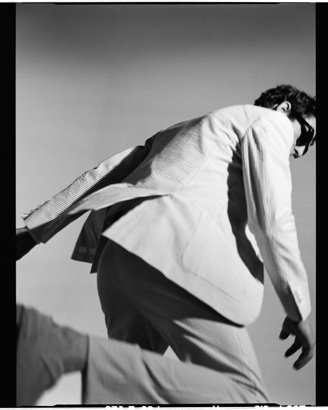 Monochromatic portrait of a man in a suit jacket and sunglasses in motion, set against a light background with soft lighting.