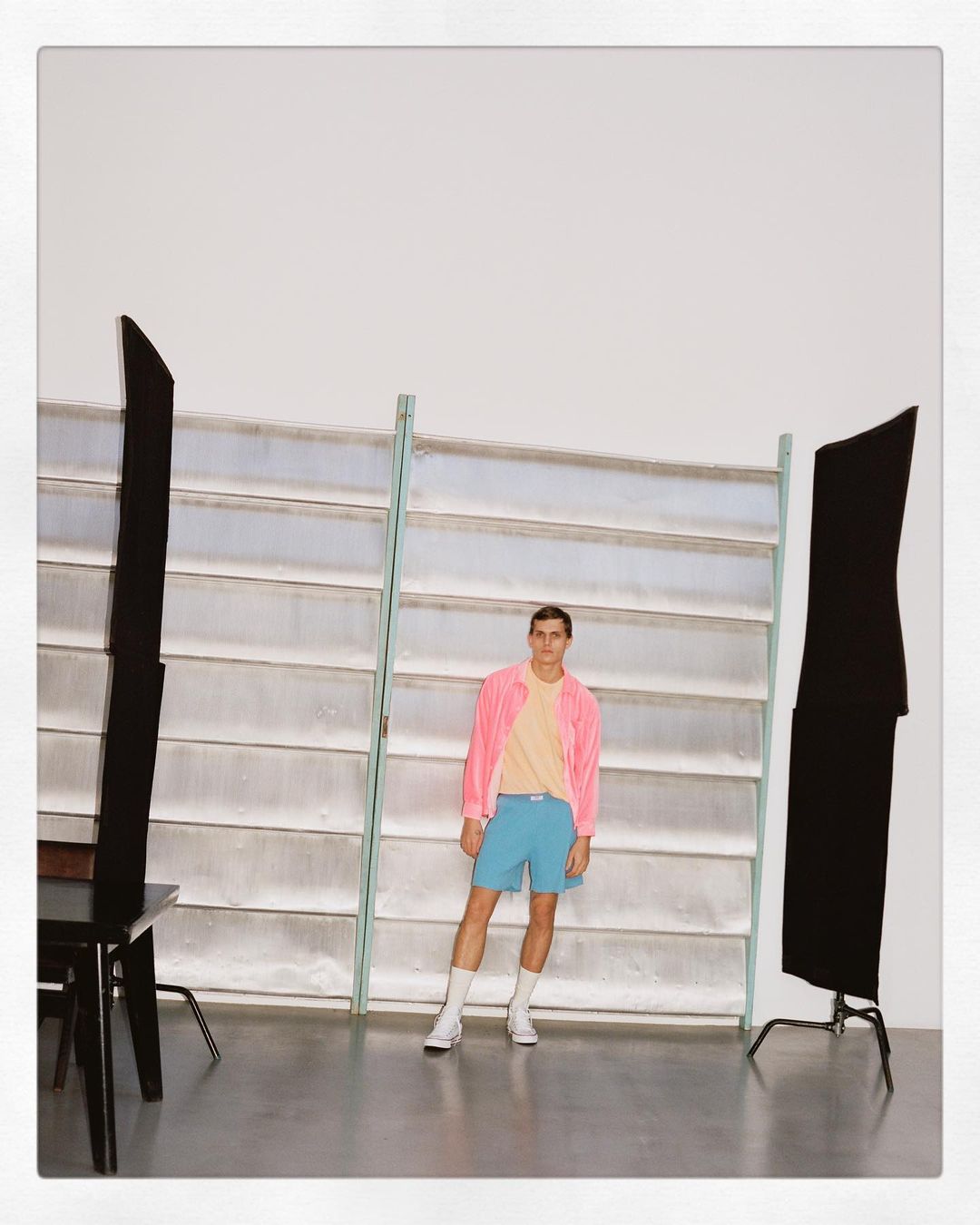 Young adult male in a pink jacket, light yellow t-shirt and blue shorts posing in a photo studio with lighting equipment.