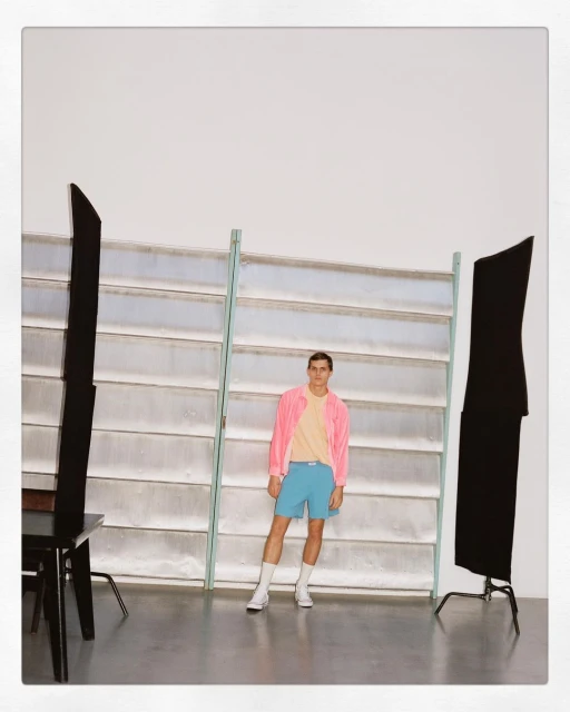 Young adult male in a pink jacket, light yellow t-shirt and blue shorts posing in a photo studio with lighting equipment.