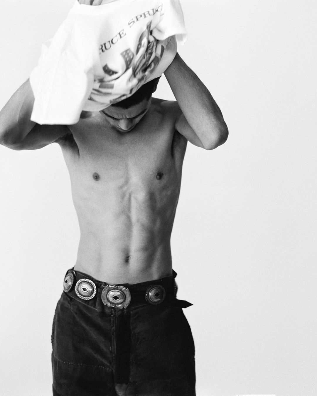 Monochrome studio shot of a shirtless male model with dark pants and silver concho belt, removing a white t-shirt with graphic print.