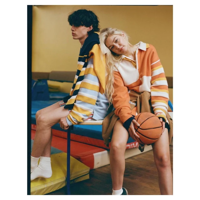 A young adult couple sits side-by-side indoors, wearing striped collared shirts. One holds a basketball in a casual lifestyle shot.