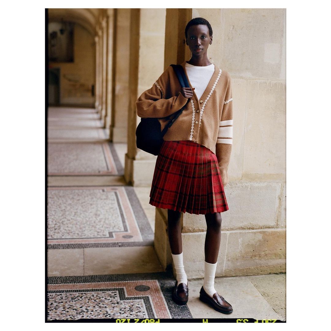 Fashion shot of an African-descent female in red plaid skirt, tan cardigan, white top, loafers, and white socks in an outdoor setting.