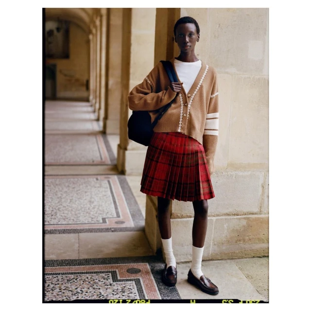 Fashion shot of an African-descent female in red plaid skirt, tan cardigan, white top, loafers, and white socks in an outdoor setting.